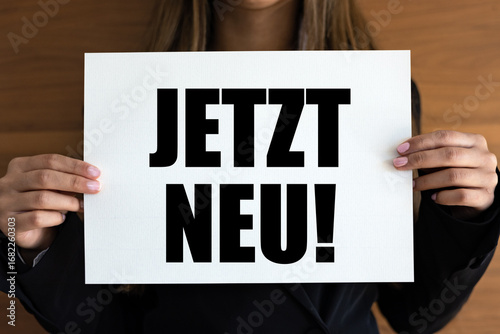 Jetzt neu! (Now new!) Woman holds a white page. Launch event, modern, inofrmation sign, premiere event, marketing.