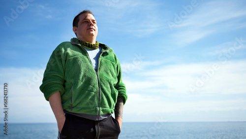 A happy, confident, obese guy in a green sweatshirt and black pants stands against a background of blue sky and sea. The man enjoys the view