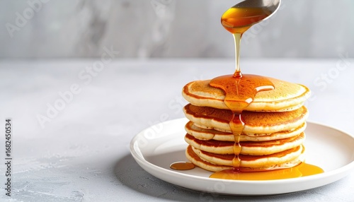 A stack of pancakes, drizzled with golden syrup from a spoon, sits on a white plate against a soft, gray background. The syrup cascades down the pancakes