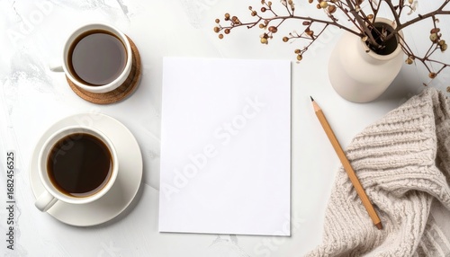 Minimalist flat lay with two coffee cups, blank paper, pencil, knitted fabric, and flower vase on a white marble background creating a cozy, inviting scene