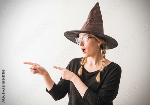 Surprised young woman in witch costume with black hat and glasses pointing sideways, isolated on white background, Halloween portrait with copy space for spooky holiday design and celebration.
