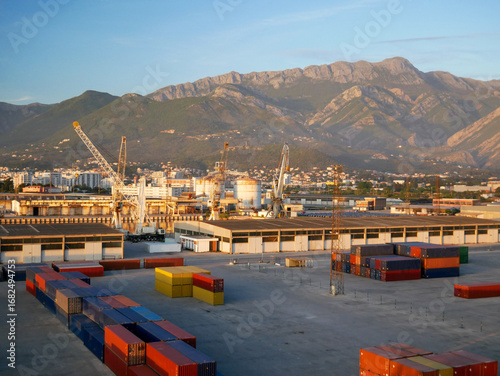 Container storage yard at the Port of Bar, Montenegro, with colorful shipping containers, cranes, warehouses and residential buildings, backed by scenic green mountains under blue sky.