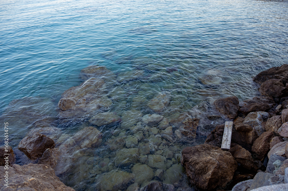 Obraz premium Close-up view of rocky coastline with transparent turquoise sea water. The photo captures the clarity of the waves gently covering the stones, creating a peaceful and refreshing seaside atmosphere.