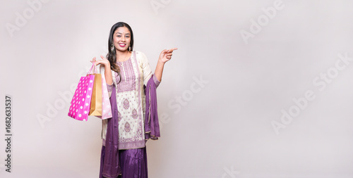 Young indian woman wearing traditional outfit holding shopping bags pointing at blank space standing isolated on white studio background.Diwali celebration and festive sale. Advertisement Copy space.