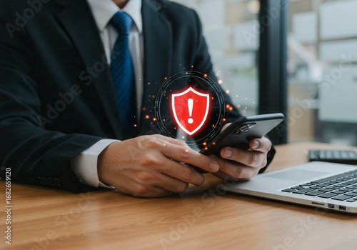A suited professional at a desk, looking at a smartphone displaying a digital warning symbol—a red shield with a white exclamation mark