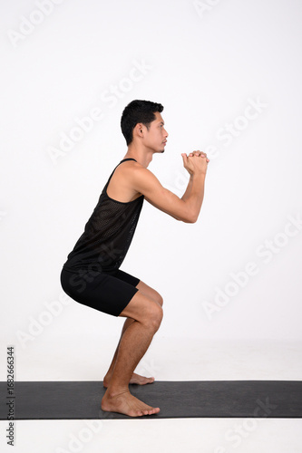 A male runner performs a bodyweight squat on a fitness mat, clasping his hands in front of his chest. Side View.