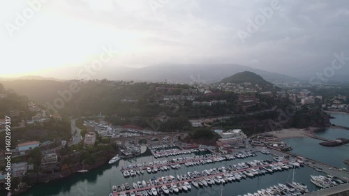 drone footage captures a marina filled with boats at Mandelieu La Napoule in the South of France. The setting includes lush hills, coastal architecture, and a glowing sunset