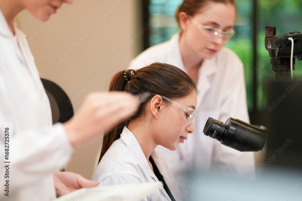 Obraz premium A team of female professionals works together in a science lab. One woman looks into a microscope while her colleagues help and discuss the results of their project.