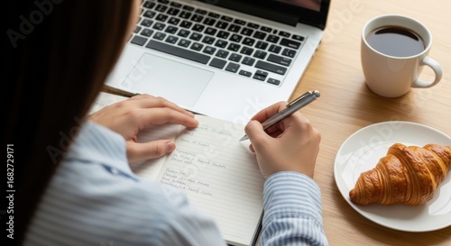 Female writing in notebook with coffee and croissant by laptop on wooden desk