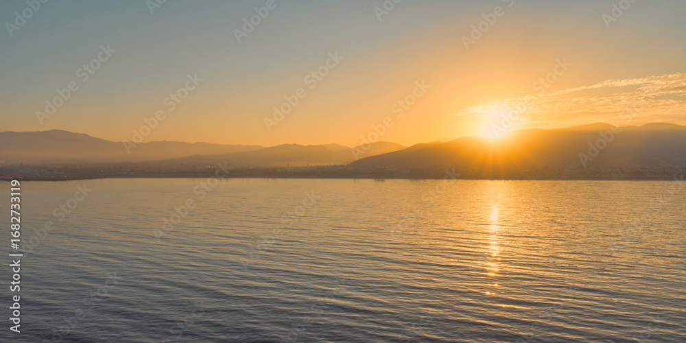 Obraz premium Peaceful sunrise over calm waters with a warm golden glow reflecting on the gentle waves and distant mountains in the background.