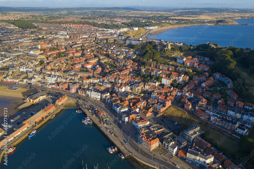 Obraz premium Aerial morning view of Scarborough town and harbour, with rows of colourful houses, fishing boats along the quayside, and sweeping views of the sandy bay and surrounding coastline.