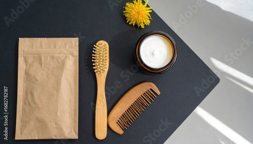 Top-down shot of natural hair & skin care items (comb, brush, cream, packet) & a yellow flower on a two-tone background, with sunlight
