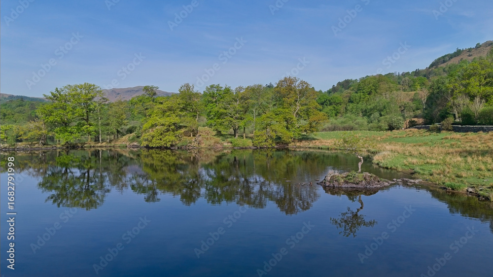 Fototapeta premium A beautiful, peaceful Rydal water lake reflects the surrounding green trees under a bright blue sky.