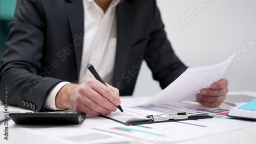 Close up of a male hand filling out documents with a pen using a calculator sitting at a desk at a workplace in a business office. Businessman is engaged in paperwork, making financial calculations