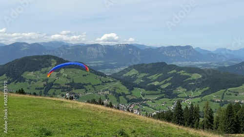Majestic view of two paragliders against a mountain landscape. A red paraglider hovers in the air, while a blue one lifts off the ground and hovers, capturing the thrill of flight and adventure