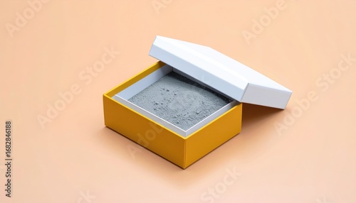 A square yellow box with an open white lid contains grey powder, set against a light peach background in a studio setting