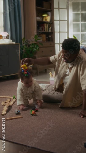 Vertical shot of young African American father watching little baby girl taking first steps at home and holding toys