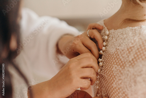 Tender bridal morning. The bride in a soft robe admires her gown, bridesmaids fastening delicate lace details. A touching pre-wedding ritual filled with elegance, calm, and graceful charm.