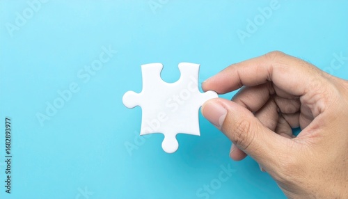 Close-up of a hand holding a single, white jigsaw puzzle piece against a light blue background, conveying problem-solving or a missing element