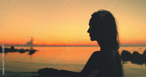 A silhouette of a woman sitting on a sandy beach, framed against the vibrant hues orange sunset in Thailand. The calm waters reflect the colorful sky as she gazes out towards the horizon.