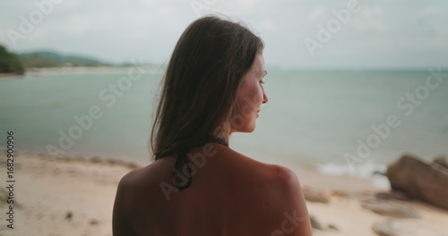 Beautiful girl relax on resort bungalow terrace on holiday summer vacation. Tropical island sea landscape empty sand beach coast in background. Travel to Thailand exotic island. Back view zoom out