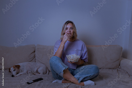 Young woman watching a movie at home with a surprised expression, holding a bowl of popcorn. Cozy evening scene, emotional reaction, casual outfit, dog sleeping nearby.