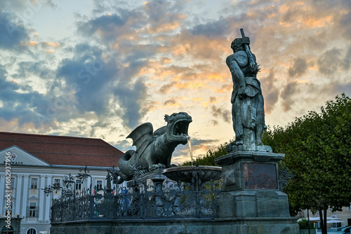 At sunset in Klagenfurt, Austria, the Lindwurmbrunnen statue stands silhouetted against the glowing sky, a symbol of history, culture, and the city’s enduring heritage and identity.