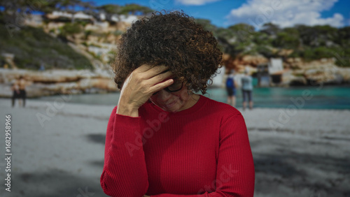 Canvas Print Woman with curly hair in red sweater and glasses folding arms with hand on forehead at sandy beach; silent discomfort