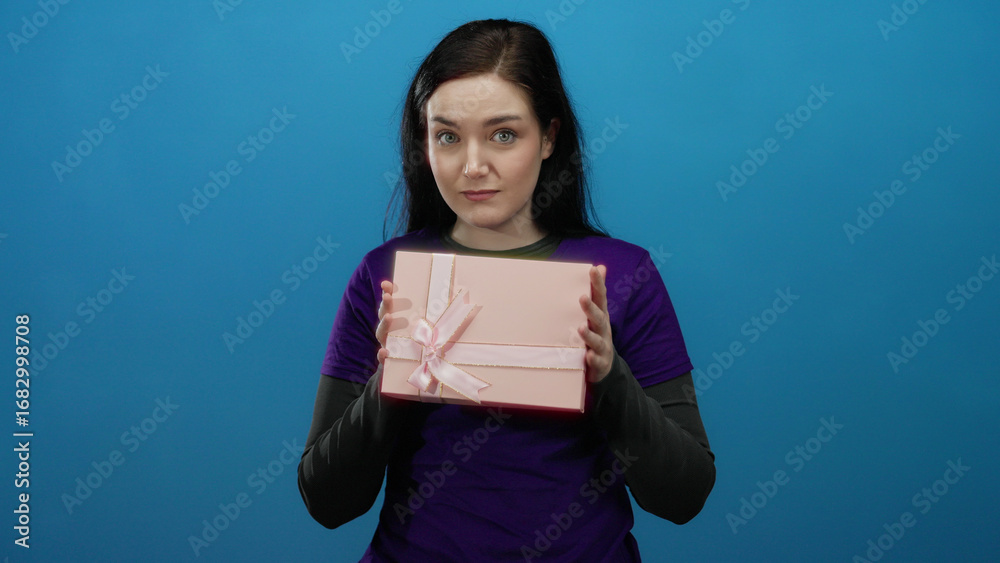 Obraz premium Woman holding pink gift box in front of blue background wearing purple volunteer shirt with joyful expression.
