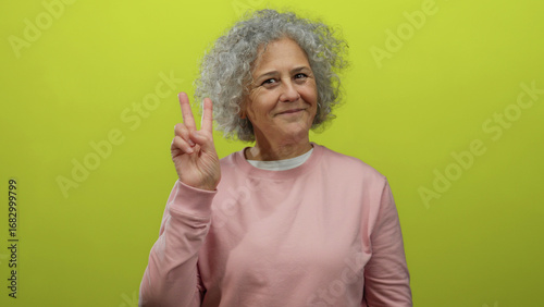 Woman with grey hair smiling and showing peace sign against a vibrant yellow background wearing a pink sweater conveying happiness and positivity.