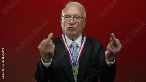 Senior man in a business suit with medal gestures rudely on an isolated red background, capturing a provocative and unorthodox senior expression.