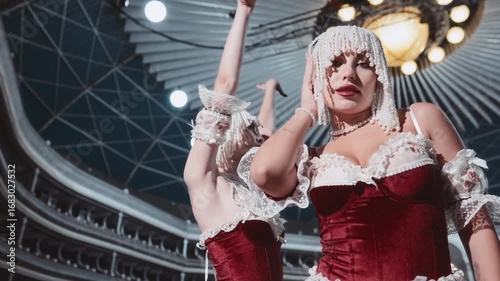 Two glamorous women in burlesque outfits pose on an empty theater stage. They are wearing red corsets with lace, pearl headdresses, and pearl necklaces.