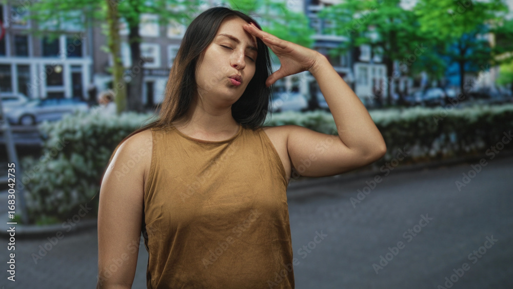 Fototapeta premium Woman hand to forehead on street in front of row houses and hedges, lips pursed gesture wearing brown sleeveless top; annoyance.