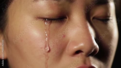 Extreme close-up shot of a young woman's face, eyes closed, with a single tear rolling slowly down her cheek. The background is completely black, and a soft, warm light illuminates only the side of he