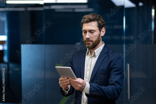 A young successful businessman in a suit is standing in an office building and using a tablet he is holding in his hands