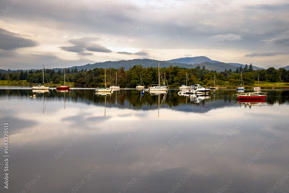 Obraz premium yachts and boats reflected in the still, calm waters of a huge lake with distant trees and mountains, Lake Windermere, Cumbria, The Lake District, England 
