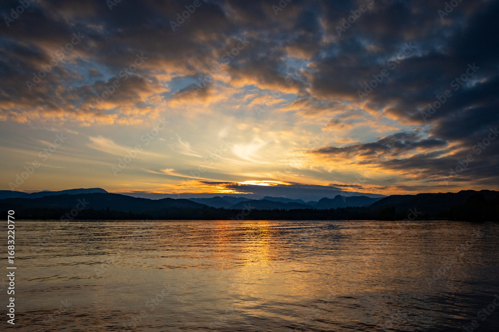 Naklejka premium Colourful cloudy sunset reflected in the ripples of a calm lake, with distant mountains, Lake Windermere, The Lake District, Cumbria, England