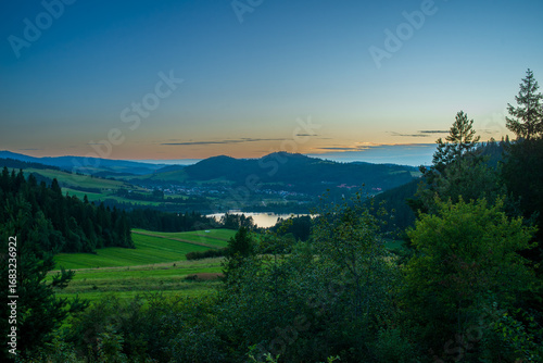 Fototapeta Naklejka Na Ścianę i Meble -  Green meadows and dark forests surround the shimmering Czorsztyn Lake as the last light of day fades. The panoramic view captures the quiet beauty of southern Poland, ideal for eco-tourism and rural t
