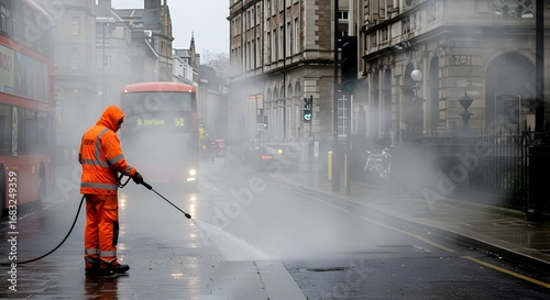 City worker in an orange high-visibility suit pressure washing a street on a foggy day with a red double-decker bus in the background