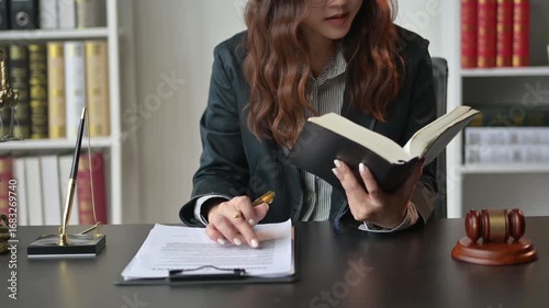 woman in a suit is reading a book while sitting at a desk. She is holding a pen and a clipboard. The scene suggests a professional setting, possibly in a law office or a courtroom