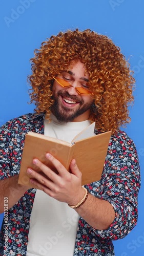 Happy relaxed smiling man with curly hairstyle wig reading funny interesting fairytale story book, leisure hobby, knowledge wisdom, education, learning, study, wow. Guy isolated on blue background