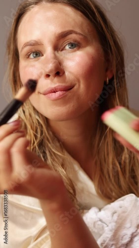 A woman is applying blush to her cheeks using a soft brush, showcasing her beauty routine. Natural light highlights her features as she enjoys the calm atmosphere in her space.