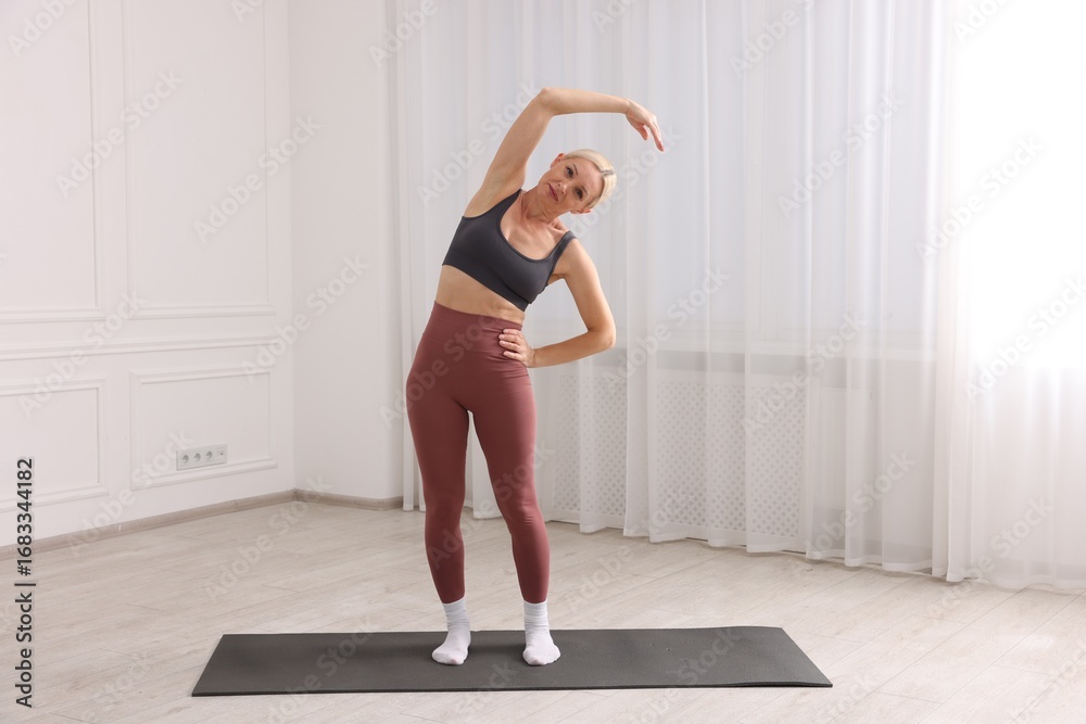 Obraz premium Mature woman in sportswear stretching on mat near window at home