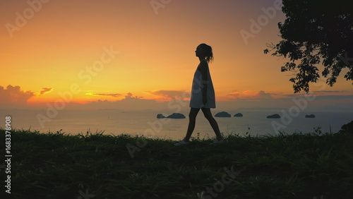 Woman gracefully walks grass hill, sunset sky in background. Female enjoy aerial view on tropical island. Girl silhouette against colorful orange sky. Outdoor lifestyle travel, summer holiday vacation