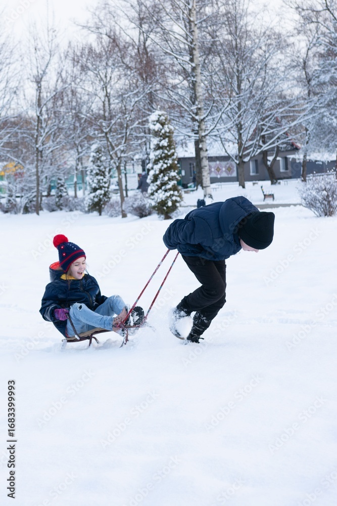 Fototapeta premium A child enjoys a snowy day being pulled on a sled by a sibling through a winter wonderland.