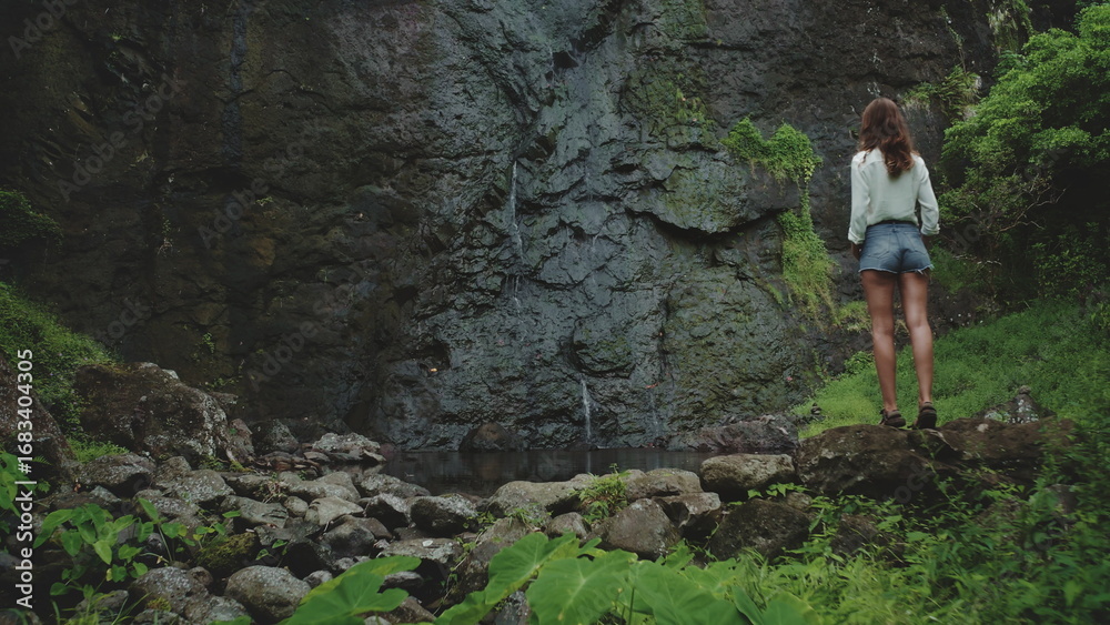 Naklejka premium Hiking woman in tropical rainforest mesmerized by waterfall on mossy rock, surrounded by lush greenery, creating peaceful moment in untouched paradise