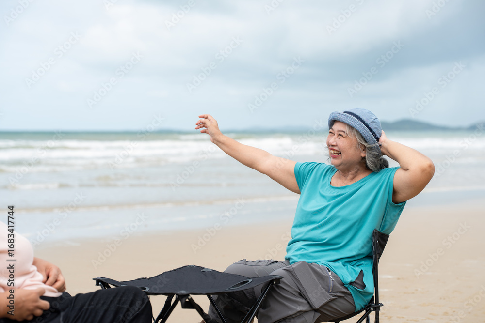 Fototapeta premium Asian senior female in casual enjoying sitting folding chair relaxing on beach seaside. Happy retired women vacation holiday lifestyle outdoor on summer.