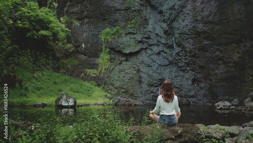 Young woman sits cross-legged on a rock, meditating in front of a lush waterfall in French Polynesia. The tropical landscape is serene and peaceful, with small pool of water. Back view, dark toning