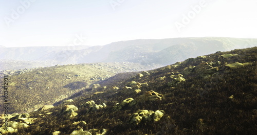 A vast and rugged landscape features rocky terrain and rolling hills under a soft morning light. The distant mountains are partially shrouded in mist, creating a serene atmosphere.