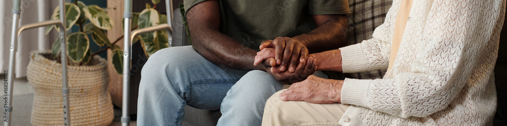 Fototapeta premium Young Black man holding hands with senior Caucasian woman sitting together indoors, showing support and compassion, partial view with walking aids and plant in background